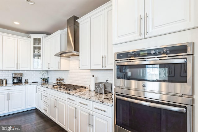 kitchen with appliances with stainless steel finishes, white cabinetry, light stone counters, decorative backsplash, and wall chimney exhaust hood