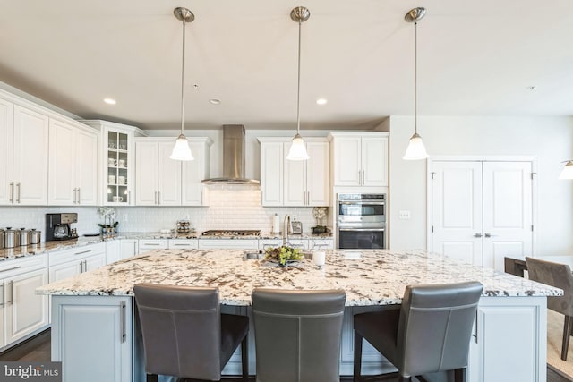 kitchen featuring a center island with sink, pendant lighting, wall chimney range hood, and white cabinets