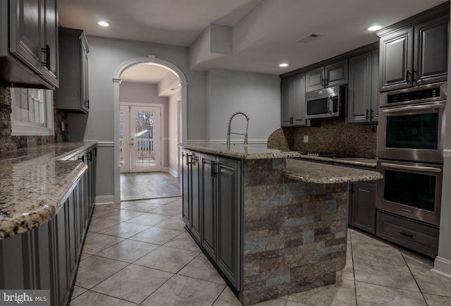 kitchen with gray cabinets, stainless steel appliances, light stone counters, tasteful backsplash, and a center island with sink