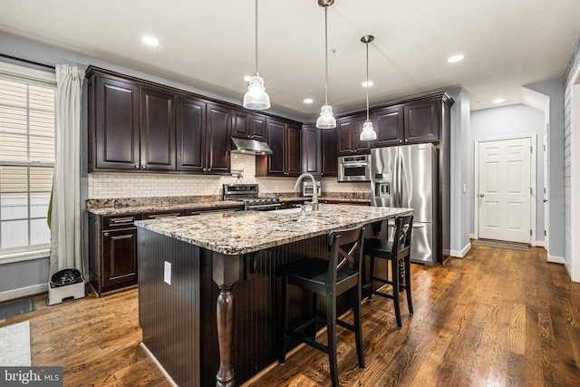 kitchen with pendant lighting, appliances with stainless steel finishes, a kitchen island with sink, dark brown cabinets, and light stone countertops