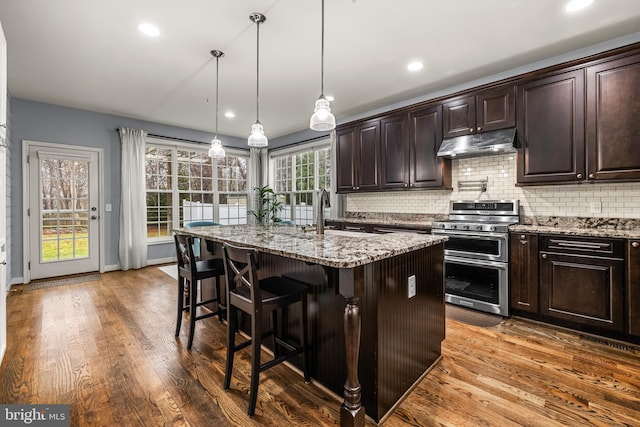 kitchen featuring a breakfast bar, light stone counters, hanging light fixtures, double oven range, and a kitchen island with sink