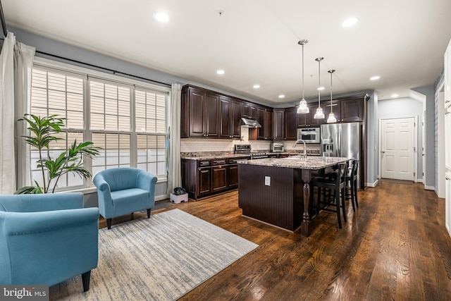 kitchen with decorative light fixtures, an island with sink, a kitchen breakfast bar, light stone counters, and dark brown cabinetry