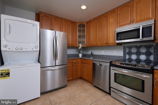 kitchen with dark stone countertops, light tile patterned floors, appliances with stainless steel finishes, stacked washing maching and dryer, and decorative backsplash