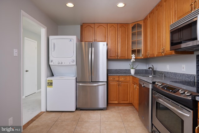 kitchen with sink, light tile patterned floors, stacked washing maching and dryer, dark stone countertops, and appliances with stainless steel finishes