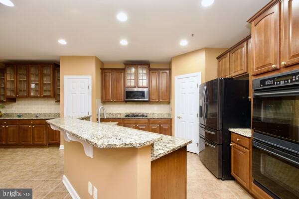 kitchen featuring a kitchen island with sink, light stone counters, tasteful backsplash, black appliances, and a kitchen bar