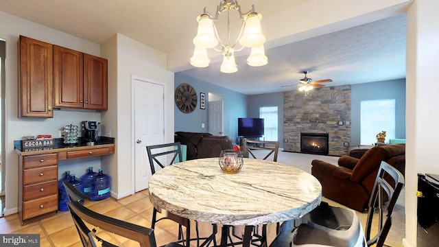 dining space with light tile patterned floors, a stone fireplace, and ceiling fan with notable chandelier