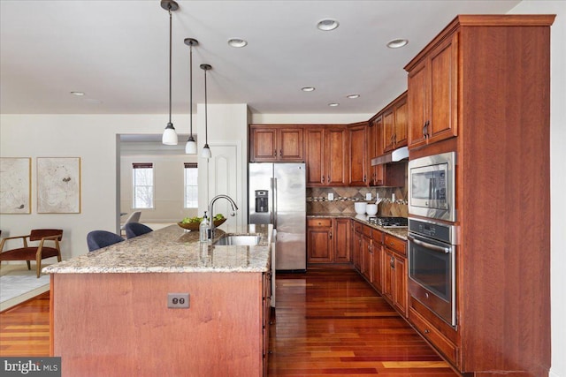 kitchen featuring an island with sink, appliances with stainless steel finishes, pendant lighting, and light stone counters