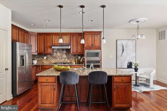 kitchen with backsplash, stainless steel appliances, light stone counters, an island with sink, and decorative light fixtures