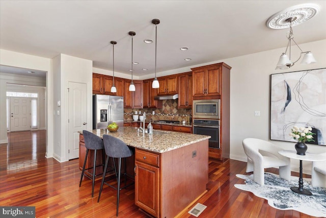 kitchen featuring tasteful backsplash, decorative light fixtures, appliances with stainless steel finishes, an island with sink, and light stone countertops