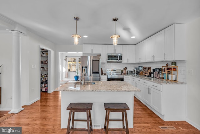 kitchen featuring appliances with stainless steel finishes, an island with sink, pendant lighting, light stone countertops, and white cabinets