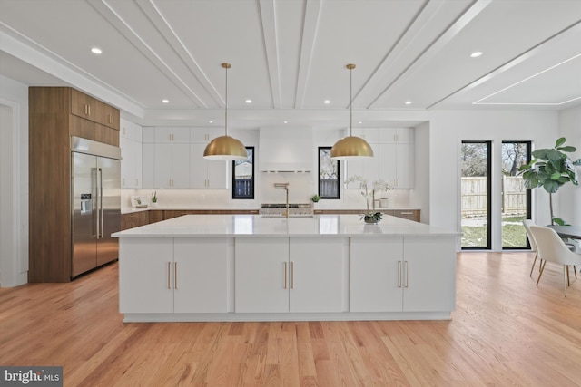 kitchen with white cabinetry, light hardwood / wood-style floors, stainless steel built in fridge, decorative light fixtures, and a large island with sink