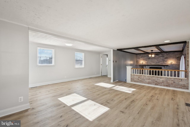 unfurnished living room featuring a brick fireplace, light wood-type flooring, beamed ceiling, ceiling fan, and brick wall