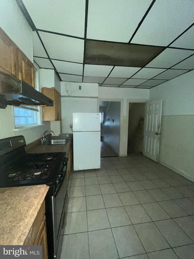 kitchen featuring sink, a paneled ceiling, black gas range, light tile patterned flooring, and white fridge