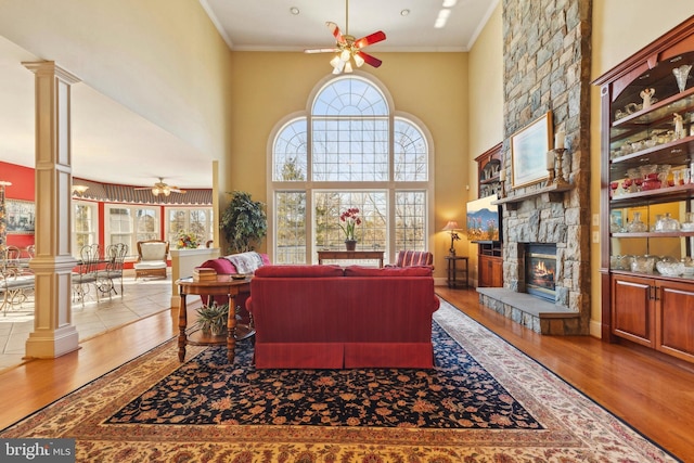 living room featuring hardwood / wood-style flooring, ceiling fan, ornamental molding, a stone fireplace, and ornate columns