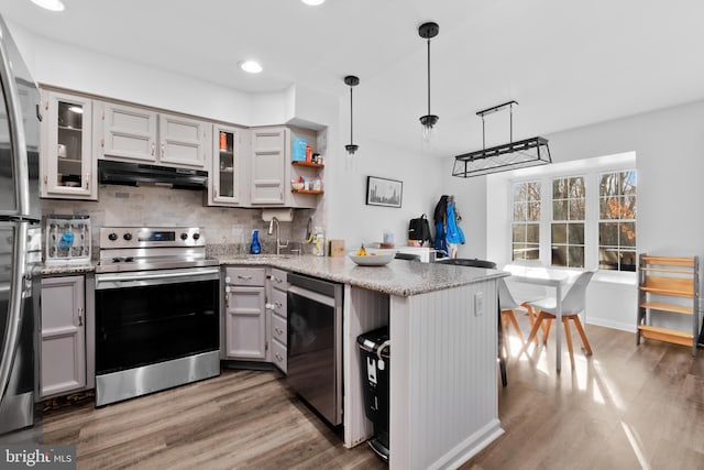 kitchen featuring pendant lighting, appliances with stainless steel finishes, light stone counters, wood-type flooring, and kitchen peninsula