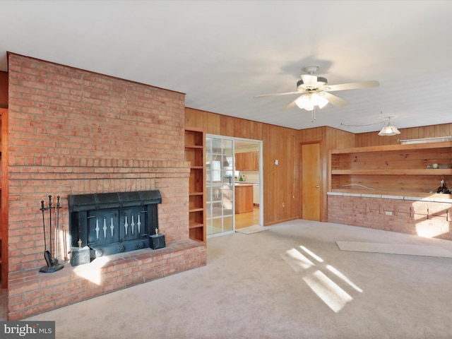 unfurnished living room featuring ceiling fan, light colored carpet, a brick fireplace, and wood walls