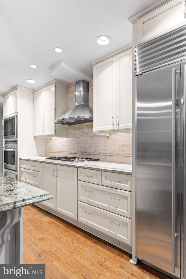 kitchen with built in appliances, light stone countertops, light wood-style floors, decorative backsplash, and wall chimney exhaust hood