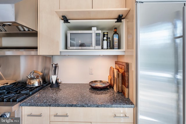 kitchen featuring stainless steel appliances, light brown cabinetry, wall chimney range hood, and dark stone counters