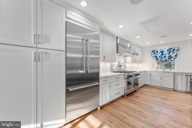 kitchen featuring white cabinets, high end appliances, light hardwood / wood-style floors, light stone counters, and wall chimney range hood