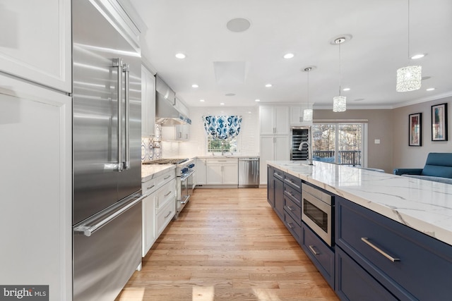 kitchen with pendant lighting, white cabinetry, built in appliances, light stone countertops, and wall chimney range hood