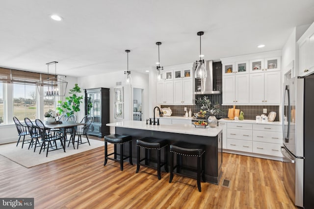 kitchen featuring wall chimney exhaust hood, a center island with sink, light hardwood / wood-style flooring, stainless steel fridge, and white cabinets