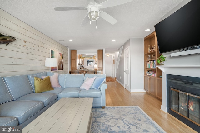 living room featuring a textured ceiling, light wood-type flooring, wooden walls, built in features, and ceiling fan