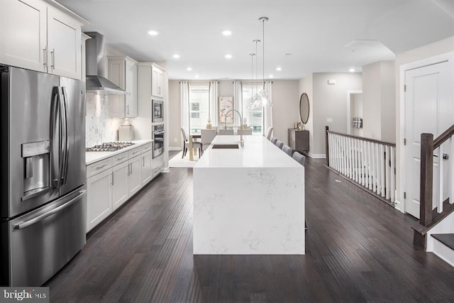 kitchen featuring sink, wall chimney range hood, appliances with stainless steel finishes, hanging light fixtures, and an island with sink