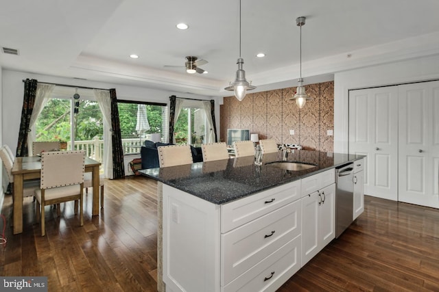 kitchen with white cabinetry, decorative light fixtures, a center island with sink, a tray ceiling, and dark stone counters