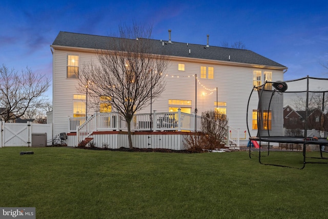 back house at dusk with a wooden deck, a trampoline, and a lawn