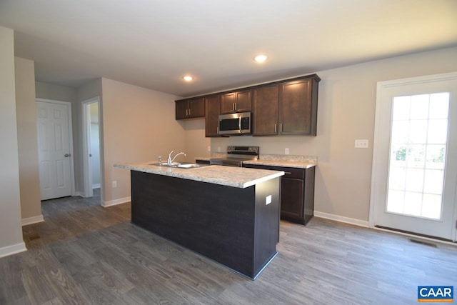 kitchen with sink, appliances with stainless steel finishes, dark hardwood / wood-style floors, dark brown cabinetry, and a center island with sink