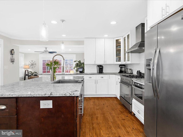 kitchen featuring wall chimney exhaust hood, sink, hanging light fixtures, appliances with stainless steel finishes, and an island with sink