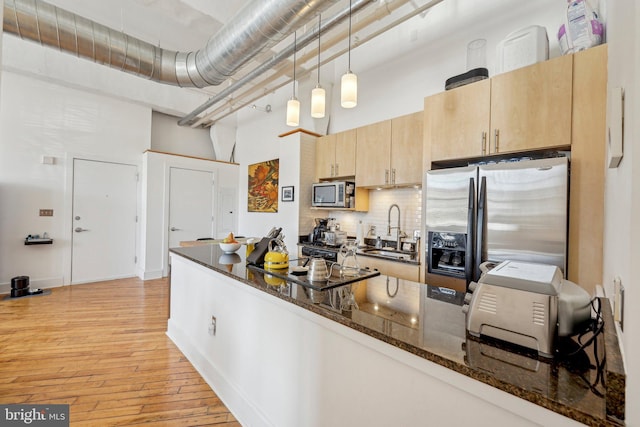 kitchen with a towering ceiling, stainless steel appliances, light brown cabinetry, decorative light fixtures, and dark stone counters