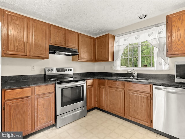 kitchen featuring appliances with stainless steel finishes, under cabinet range hood, a sink, light floors, and brown cabinetry
