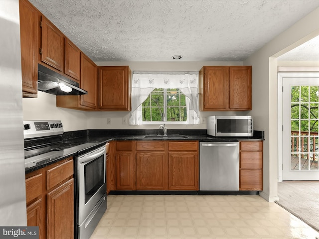 kitchen with stainless steel appliances, under cabinet range hood, a sink, light flooring, and brown cabinetry