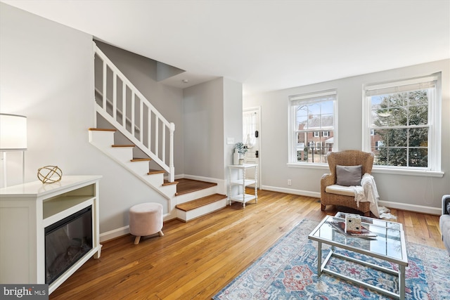 living area featuring a glass covered fireplace, stairway, baseboards, and wood finished floors