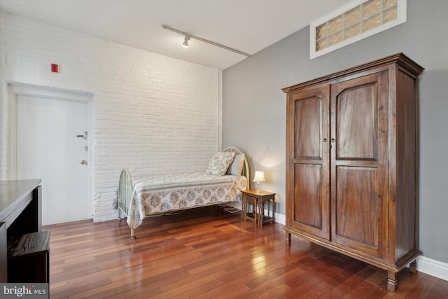 bedroom with dark wood-type flooring, rail lighting, and brick wall