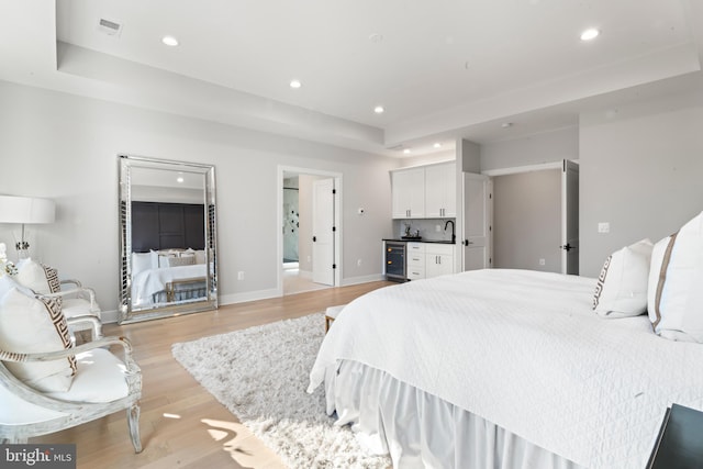 bedroom with a raised ceiling, ensuite bathroom, beverage cooler, and light wood-type flooring