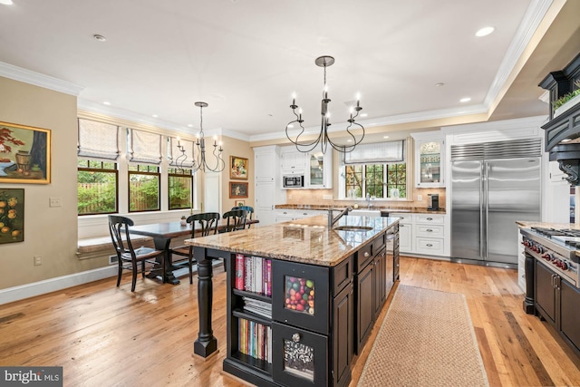 kitchen with white cabinetry, light stone counters, built in appliances, a chandelier, and a center island with sink