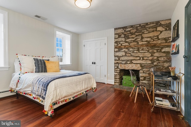 bedroom with dark wood-type flooring, a baseboard radiator, a stone fireplace, and a closet