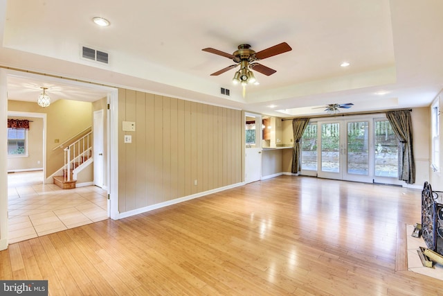 unfurnished living room featuring french doors, wood walls, a raised ceiling, ceiling fan, and light hardwood / wood-style floors