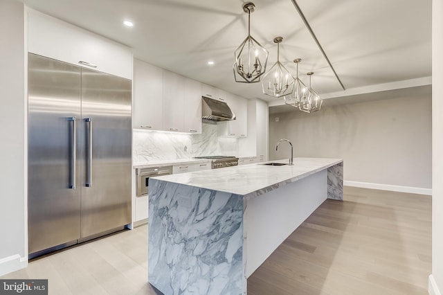 kitchen with appliances with stainless steel finishes, white cabinetry, light stone countertops, a center island with sink, and decorative light fixtures