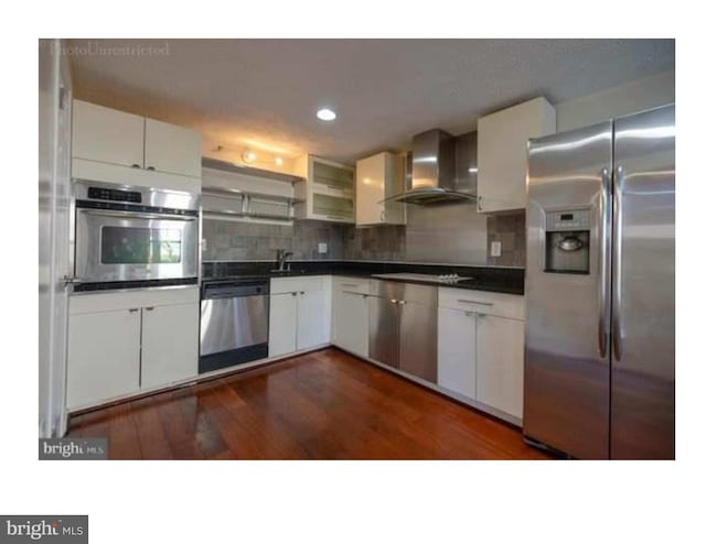 kitchen with dark wood-type flooring, stainless steel appliances, tasteful backsplash, white cabinets, and wall chimney exhaust hood