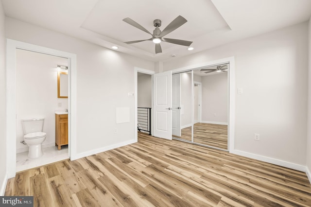 unfurnished bedroom featuring connected bathroom, light hardwood / wood-style flooring, a tray ceiling, a closet, and ceiling fan