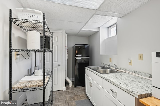 kitchen with sink, white cabinetry, black refrigerator, dark parquet floors, and a drop ceiling