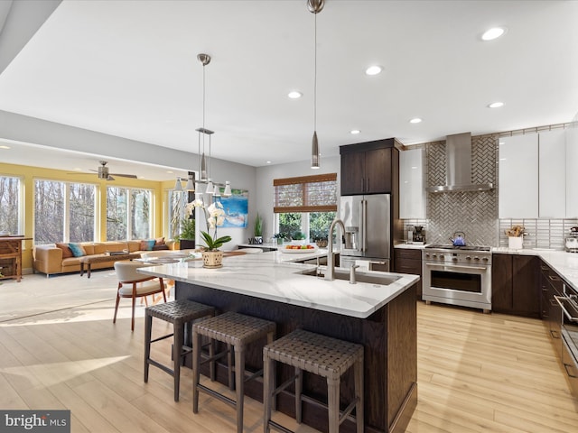 kitchen with decorative light fixtures, dark brown cabinets, high quality appliances, an island with sink, and wall chimney range hood