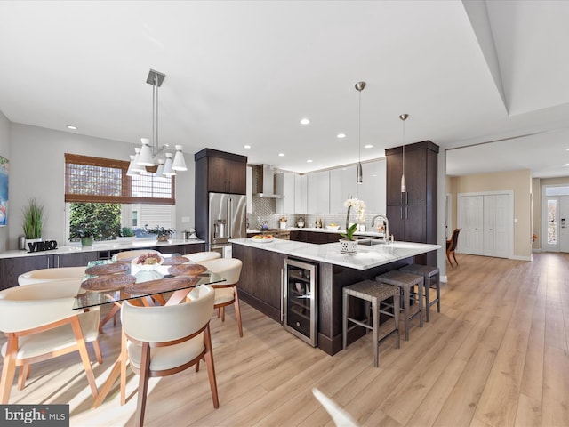 kitchen featuring pendant lighting, a kitchen island with sink, dark brown cabinets, beverage cooler, and wall chimney exhaust hood