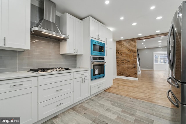 kitchen with light hardwood / wood-style flooring, wall chimney range hood, appliances with stainless steel finishes, brick wall, and white cabinets