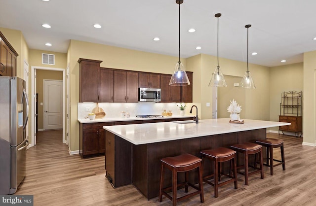 kitchen with sink, hanging light fixtures, a kitchen island with sink, stainless steel appliances, and light wood-type flooring