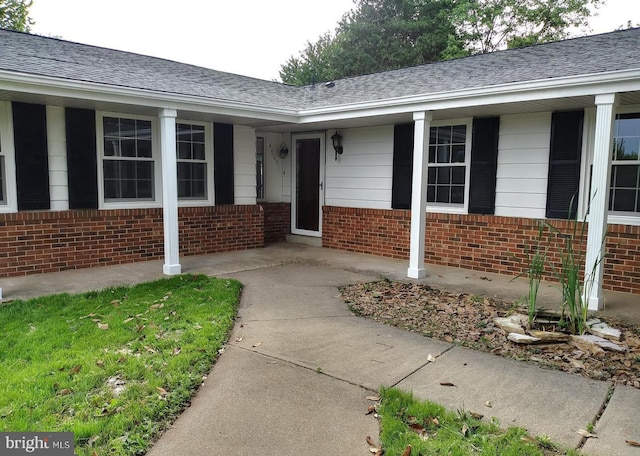 entrance to property with brick siding, covered porch, and a shingled roof