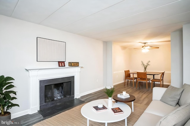 living area with dark wood-style floors, baseboards, a fireplace with raised hearth, and a ceiling fan